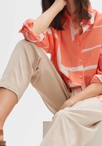 Chemise à boutons en corail avec des rayures abstraites blanches, manches retroussées, associée à un pantalon cropped beige clair. Tissu lisse et léger.