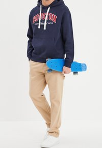 Felpa navy con la scritta "HOUSTON", pantaloni beige e sneakers bianche. Tiene uno skateboard di plastica blu. Texture lisce e design sportivo.