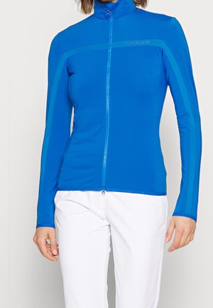 Mujer con chaqueta deportiva ajustada azul con cremallera y mangas largas, y pantalones blancos, posando frente a un fondo liso.