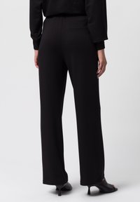 Pantalones de pierna ancha negros con una textura suave, cinturilla elástica y un discreto detalle de bolsillo en la parte trasera. Combinados con sandalias negras de tacón.