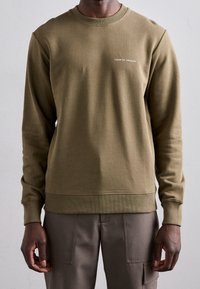 Man med en olivgrön tröja med rund hals och texten "TIGER OF SWEDEN" på bröstet, kombinerad med bruna byxor med framfickor.