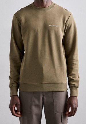 Man wearing olive green crewneck sweatshirt with "TIGER OF SWEDEN" text on chest, paired with brown pants with front patch pockets.
