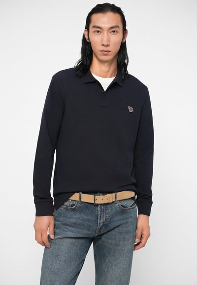 Camisa polo de manga longa azul marinho com colarinho, apresentando um logótipo bordado. Usada com jeans de tom azul claro e um cinto bege. Textura suave.