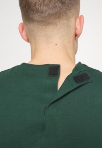 Zelené tričko s viditelným zadním otvorem zajištěným černými suchými zipy pro snadné oblékání. Měkká texturovaná látka.