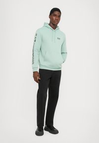 Mint green hoodie with kangaroo pocket, drawstring hood, and black text detailing on sleeve. Paired with black pants and shoes.
