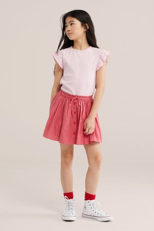 Jong meisje staat, draagt een lichtroze mouwloos topje met ruches aan de mouwen, een roodgestreepte rok, rode sokken en witte sneakers.