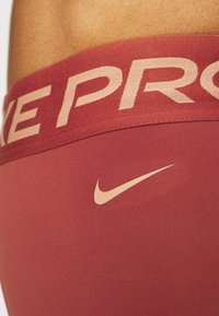 Červené sportovní legíny s širokým elastickým pasem, na kterém je nápis "NIKE PRO" v světle béžové barvě. Malé béžové logo Nike na přední straně. Hladká textura.
