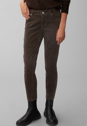 Manos descansando junto a las caderas, vistiendo pantalones de pana ajustados de color marrón oscuro combinados con botas de cuero negras y gruesas hasta el tobillo.