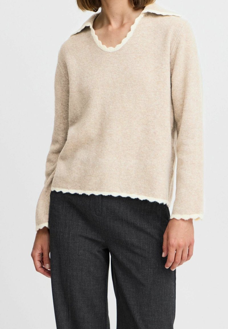 Pull beige en maille avec un col et un ourlet blancs à bord festonné, associé à un pantalon gris foncé. Le pull présente une coupe décontractée et des manches longues.