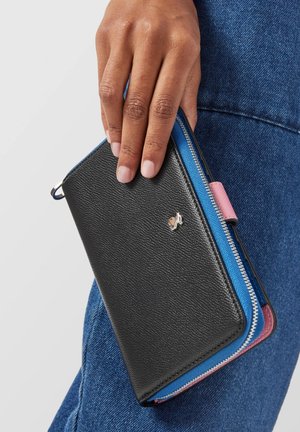 Portefeuille en cuir noir avec une surface texturée, présentant des accents bleus et roses ainsi que des fermetures éclair en métal argenté. Tenue dans une main contre un tissu en denim.
