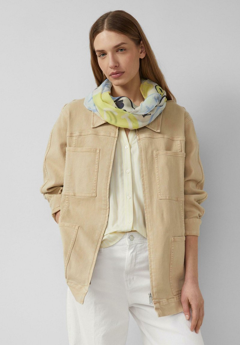 Donna che indossa una giacca beige, una camicia bianca, pantaloni bianchi e una sciarpa multicolore con tonalità gialle e blu intorno al collo.