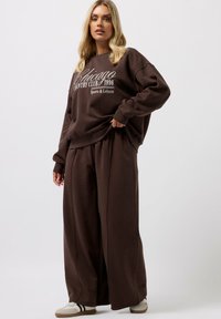 Brauner, übergroßer Sweatshirt mit weißem Textgrafik und passenden weit geschnittenen Hosen. Weicher Stoff, lockere Passform und lässiger Stil.