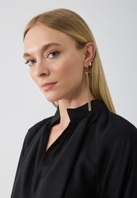 Gold statement earrings with a long tassel design, worn with a black blouse featuring a high neckline and pleated details.