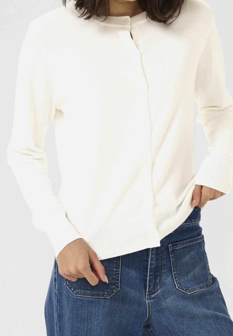 Femme portant un cardigan blanc boutonné et un jean bleu, une main dans la poche et l'autre ajustant l'ourlet du cardigan.