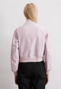 Hellrosa Bomberjacke mit geripptem Kragen und Bündchen, die eine glatte Textur aufweist. Über schwarzen Hosen getragen und damit ihr verkürztes Design zur Schau stellend.