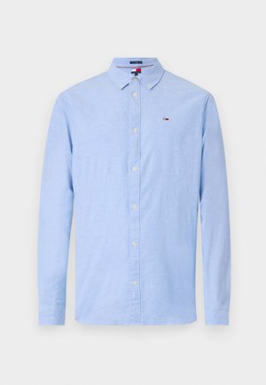 Camisa azul clara de botões, com colarinho, mangas longas e textura subtil; apresenta um pequeno logótipo na área do peito esquerdo.