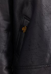 Schwarze strukturierte Lederjacke Nahaufnahme zeigt eine Tasche mit einem kleinen schwarz-gelben Alpha Industries Etikett.