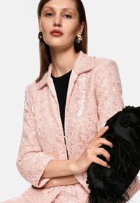 Blush-pink Spitzenblazer mit Pailletten und Reverskragen. Hält eine schwarze Clutch mit floralen Verzierungen und Federn.