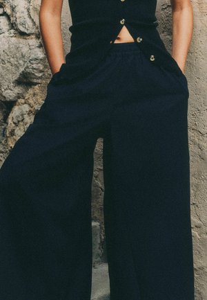 Person wearing black buttoned sleeveless top and loose black pants with hands in pockets against stone wall background.