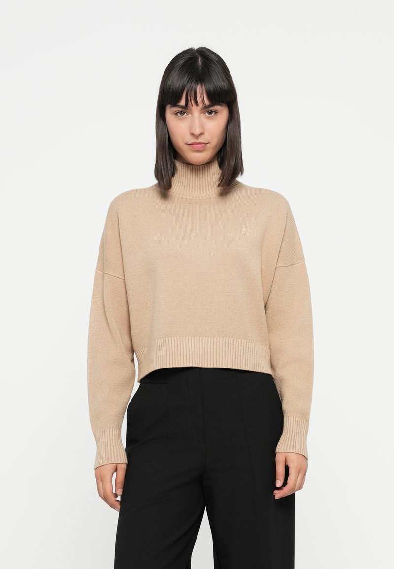 Flauschiger, gestrickter, sandfarbener Pullover mit hohem Kragen, gerippten Bündchen und verkürztem Saum. Mit minimalem Branding auf der Brust. Kombiniert mit schwarzen Hosen.