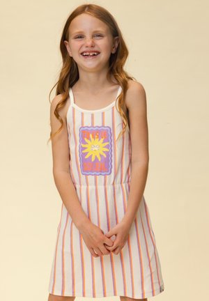 Lächelndes rothaariges Mädchen mit fehlenden Vorderzähnen, das ein weißes Kleid mit orangefarbenen und violetten Streifen und einer Sonne mit der Aufschrift "Tu es le soleil." trägt.