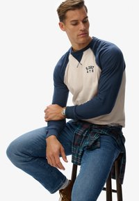 Sweatshirt à manches raglan crème et marine, portant le texte "S.DRY ATH." Associé à un jean bleu et une chemise à carreaux nouée à la taille.