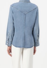 Vue de dos d'une personne portant une chemise en jean bleu clair avec un empiècement en forme de V et un pantalon blanc, sur un fond uni.