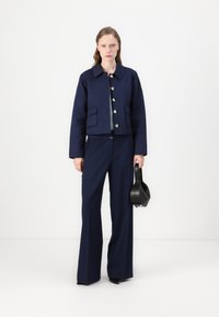 Marineblauwe cropped jacket met gouden knopen, twee voorzakken en wijde marineblauwe broek. Een zwarte handtas maakt de outfit compleet.