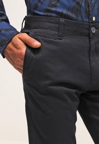 Ręka w kieszeni ciemnogranatowych spodni noszonych z niebieską, wzorzystą koszulą z długim rękawem, pokazując obszar talii i górnej części uda.