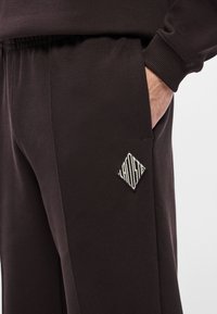 Tmavohnedé tepláky z textúrovaného materiálu, s elastickým pásom, bočnými vreckami a bielou nášivkou s logom v tvare diamantu na vrecku.