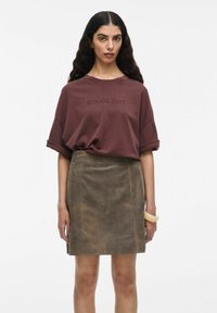 Bordo arba oversized marškiniai su užrašu "ROUGE EDIT", derinami su rudos zomšinės mini sijonu. Modelis dėvi skaidrų apyrankę ant vienos rankos.
