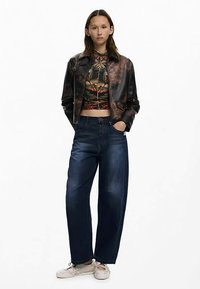 Veste en cuir noir au design court, un crop top à motifs en dessous, et un jean denim bleu foncé à jambes larges. Des baskets beiges complètent la tenue.