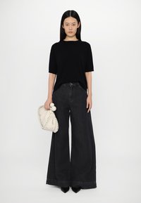 Black short-sleeve shirt paired with wide-leg dark denim jeans. Model holds a cream clutch with a braided trim and two spherical accents.