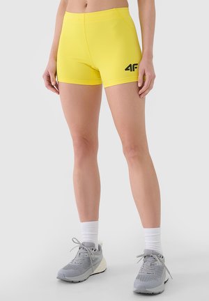 Kobieta ubrana w jaskrawożółte dopasowane spodenki sportowe z czarnym logo „4F”, białe skarpety sportowe i szare buty do biegania stoi na białym tle.