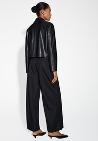 Zwarte cropped leren jas met een zachte textuur, gecombineerd met loszittende zwarte broek en puntige zwarte hakken, gezien vanaf de achterkant.