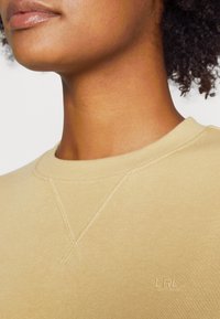 Béžová mikina s kulatým výstřihem, viditelnou ribovanou texturou a vyšívaným logem "LRL" na hrudi. Švy a měkká látka.