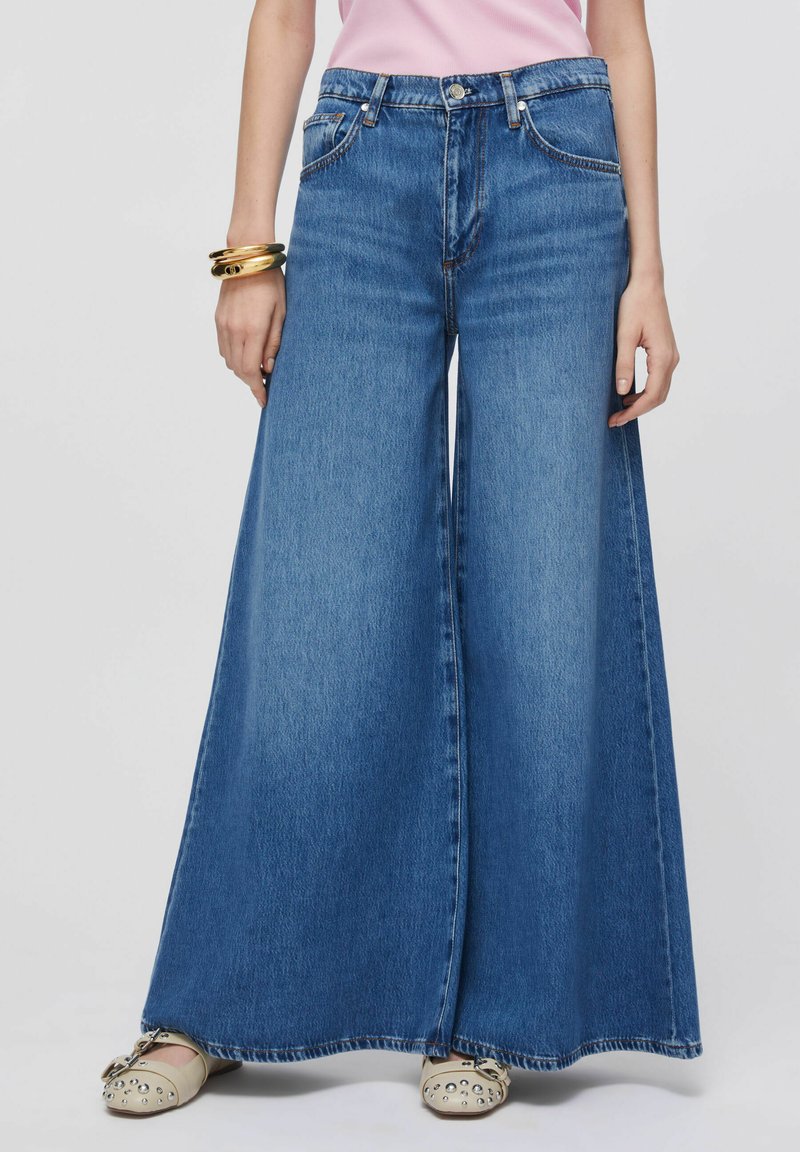 Jean en denim bleu clair à jambes larges, doté d'une taille haute, d'un design à cinq poches et d'un ourlet effiloché. Associé à des ballerines beige ornées de détails.