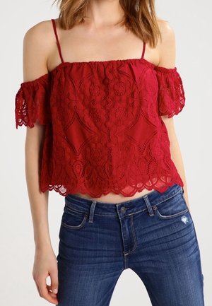 Kobieta ubrana w czerwony koronkowy top z odkrytymi ramionami i cienkimi ramiączkami, zestawiony z ciemnoniebieskimi dżinsami.