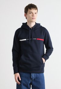 Jūros mėlynos spalvos džemperis su raišteliu ir priekiu kišene. Yra balta ir raudona horizontalinė juostelė su užrašu „TOMMY HILFIGER“ centre.