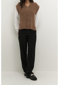 Persoon draagt een bruin mouwloos gebreid vest over een lang wit overhemd, zwarte op maat gemaakte broek en zwarte loafers met gouden detail.