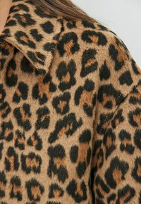 Nærbillede af blødt leopardprintstof med sorte og brune pletter på en tan baggrund, der viser detaljer om kraven og skuldersømmen.