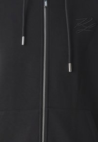 Černá mikina s plným zipem, se šňůrkami, předními kapsami a vyšitými detaily. Vyrobeno z měkkého materiálu s hladkou texturou.