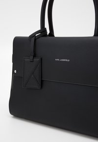 Black leather handbag with dual handles, flat design, and a textured tag. Features silver hardware and a embossed brand logo.