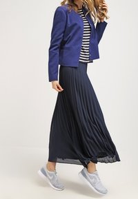 Granatowa plisowana spódnica, granatowy blezer na czarno-białej paskowanej bluzce oraz jasnoszare sportowe buty o teksturowanym wzorze.