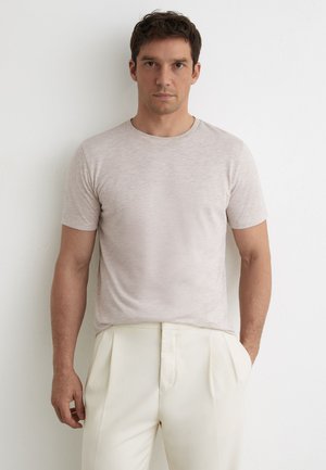 Uomo con capelli corti e scuri che indossa una camicia beige chiaro a maniche corte e pantaloni color crema, in piedi contro un muro bianco.