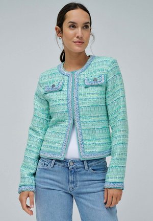 Chaqueta de tweed verde menta con patrón texturizado, ribete azul, escote redondo y dos bolsillos frontales. Combinada con jeans azul claro.