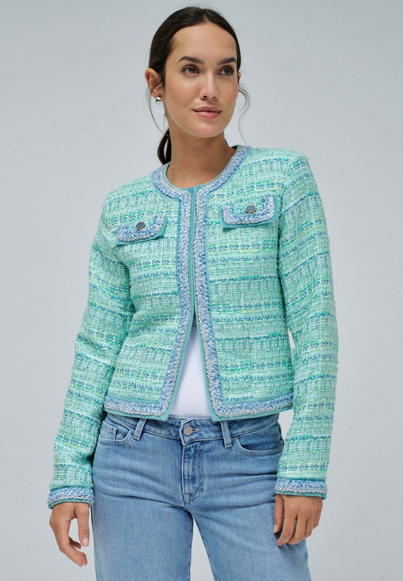 Giacca in tweed verde menta con motivo testurizzato, rifiniture blu, scollo rotondo e due tasche frontali. Abbinata a jeans azzurri chiari.