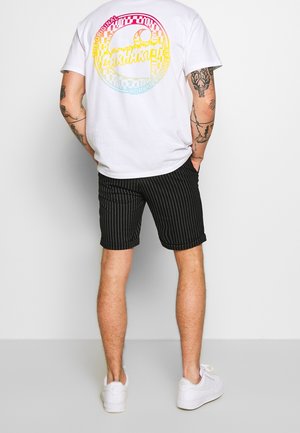 Camiseta blanca con un gráfico multicolor en la parte de atrás, combinada con pantalones cortos negros a rayas y zapatillas blancas, mostrando un atuendo casual desde la parte trasera.