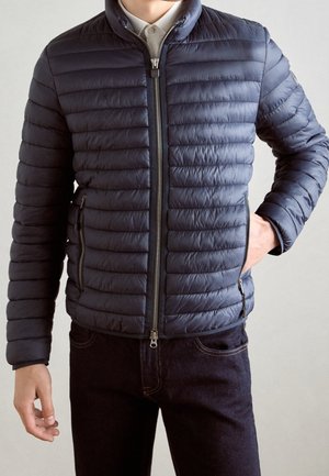 Homme portant une veste matelassée bleu marine zippée par-dessus une chemise blanche à col, avec une main dans la poche et un pantalon sombre.