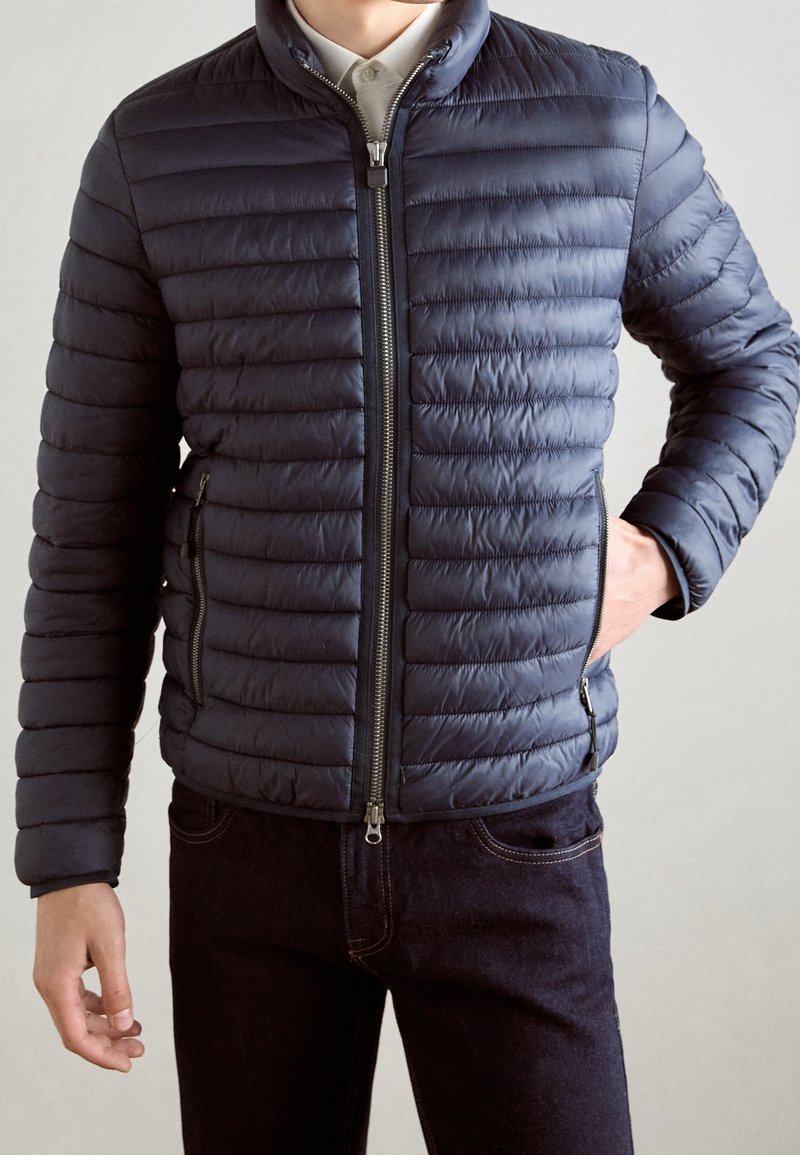 Homme portant une veste matelassée bleu marine zippée par-dessus une chemise blanche à col, avec une main dans la poche et un pantalon sombre.